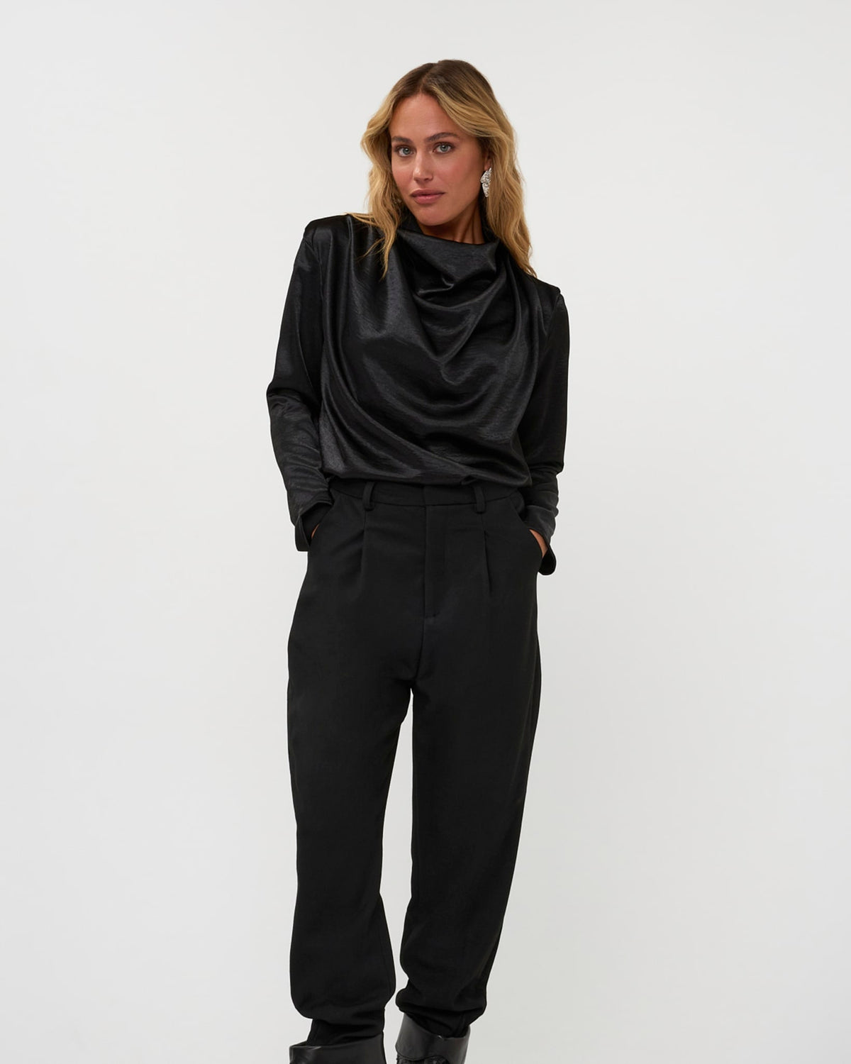 A model poses in a black cowl-neck satin top and pleated trousers, reference 264-15-1-3-18.